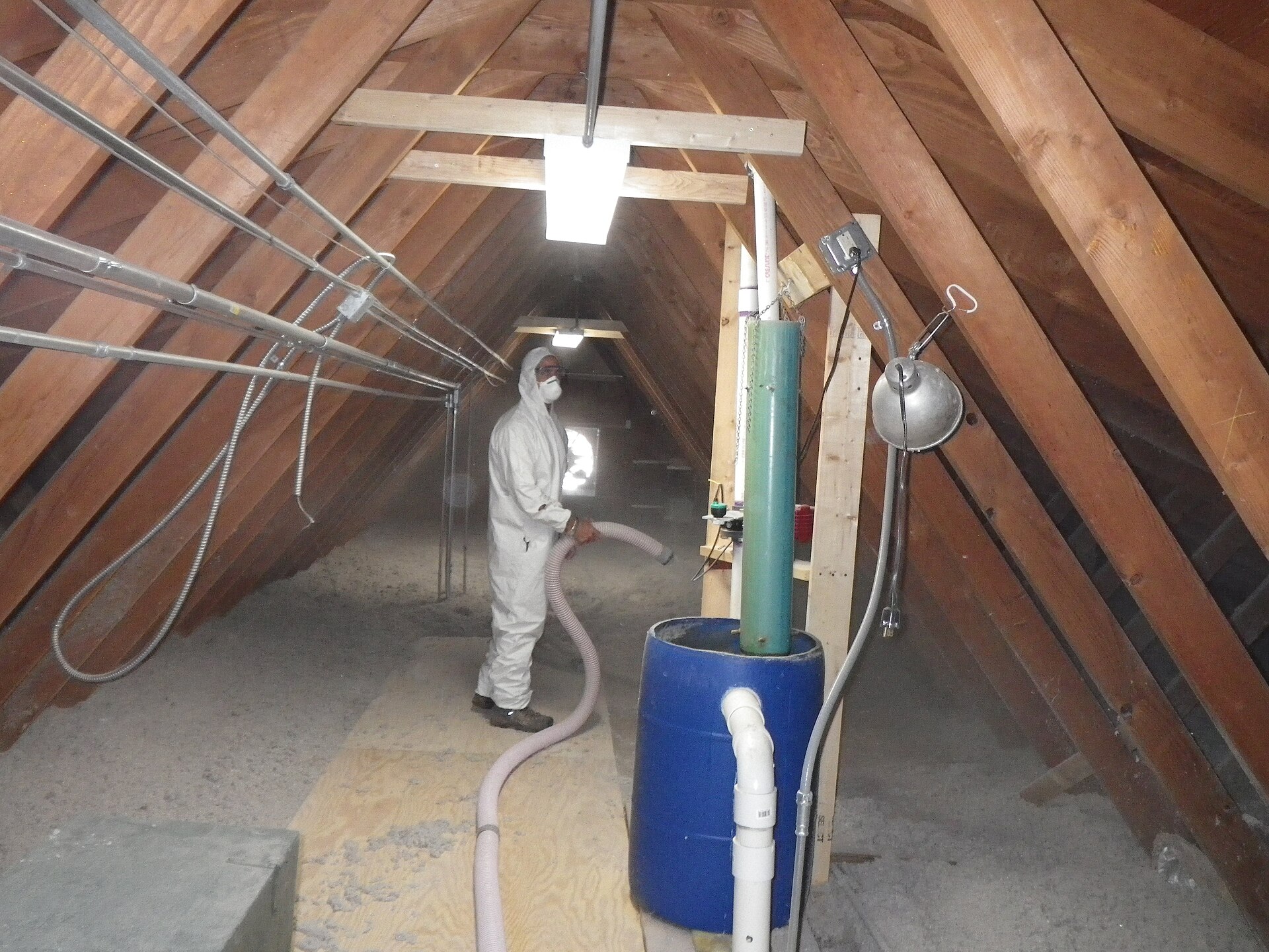 A contractor in a white protective suit uses a hose to blow gray loose-fill insulation onto an unfinished attic floor.