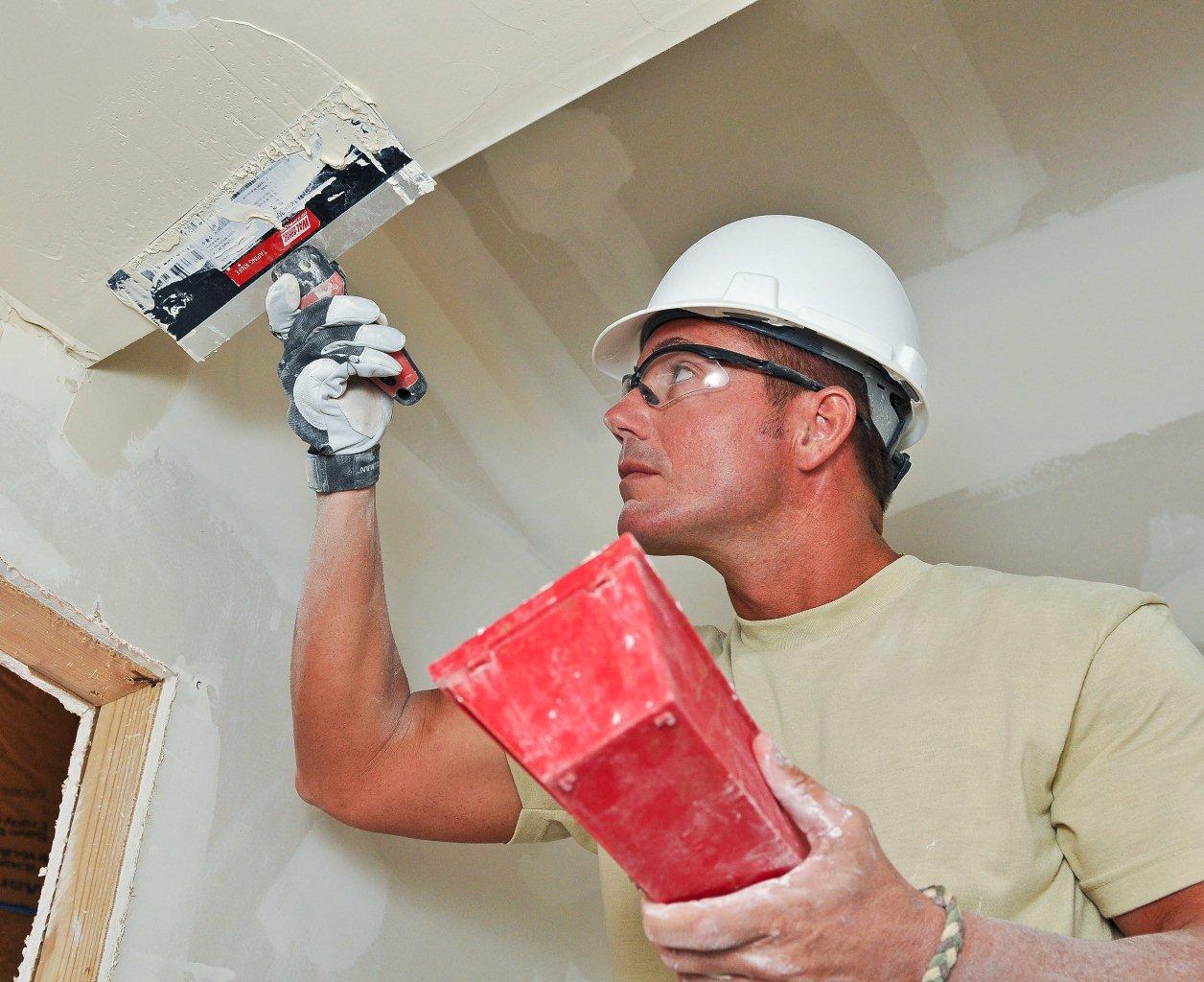 A worker in a hard hat spreading joint compound along a wall-ceiling joint with a taping knife and a red mud pan.