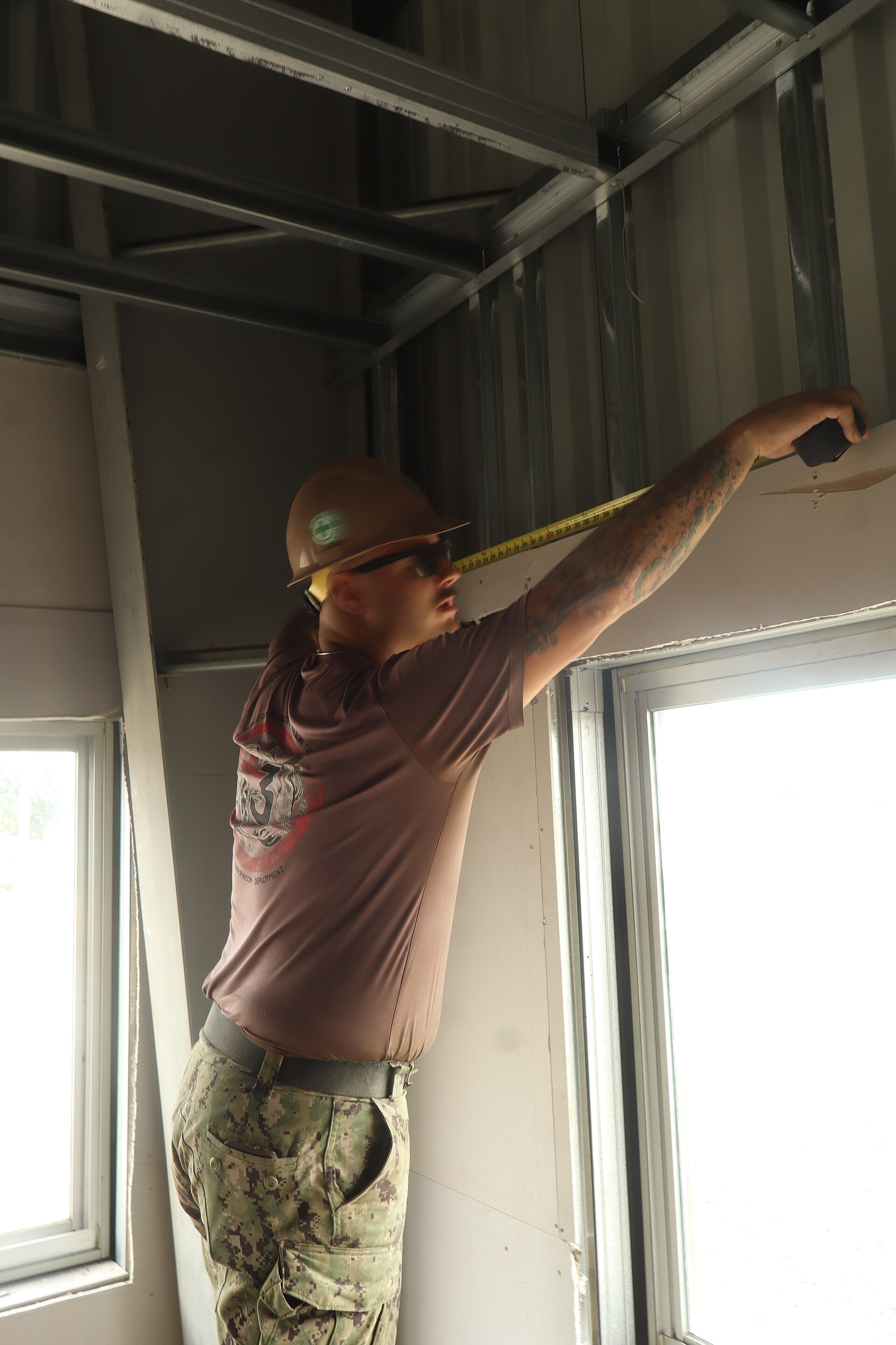A construction worker in a hard hat measuring installed drywall around a bright window opening.