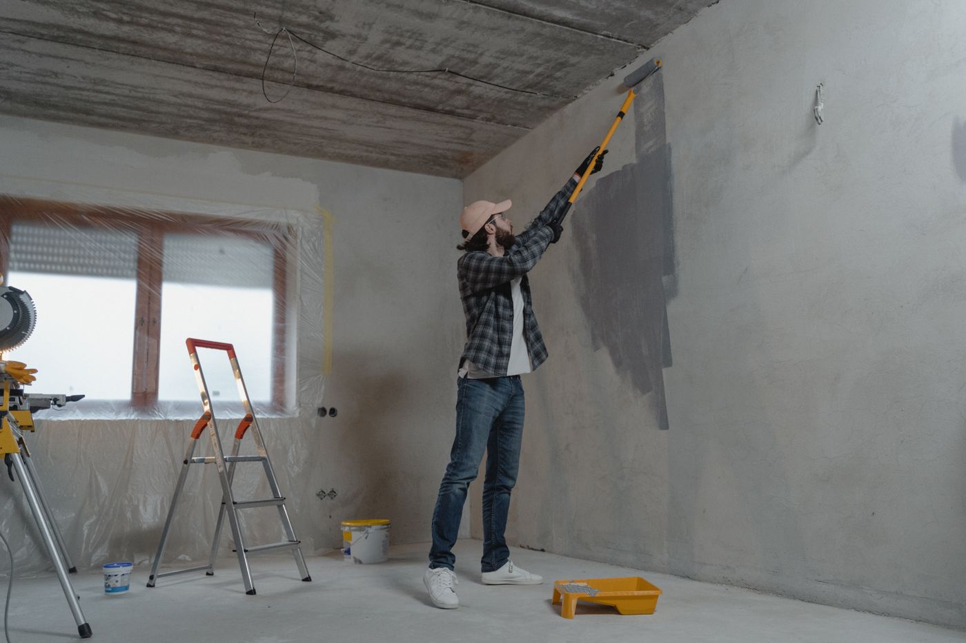 A man in a plaid shirt and baseball cap uses a roller on a yellow extension pole to apply dark gray paint to a primed interior wall.