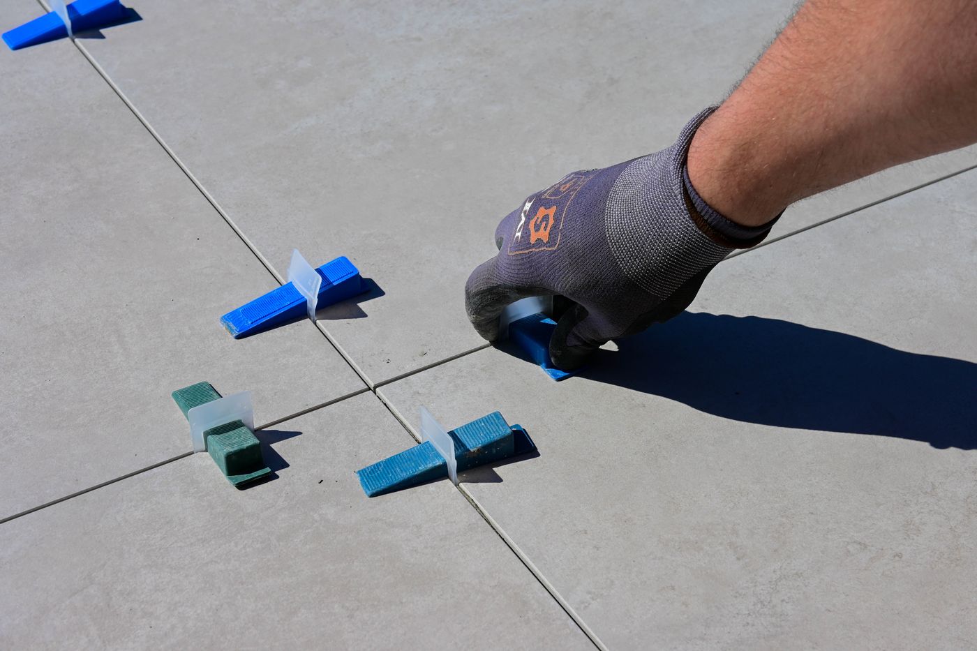 A gloved hand adjusting a blue plastic leveling wedge between light-gray floor tiles.